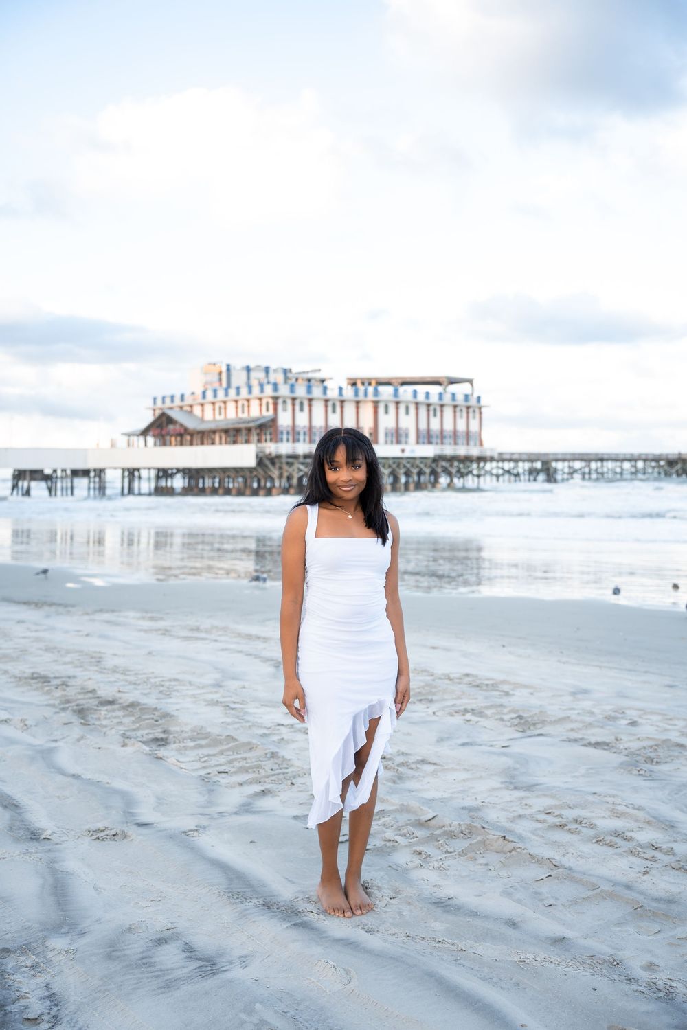 A young woman in a white dress stands barefoot on a sandy beach with a pier in the background.