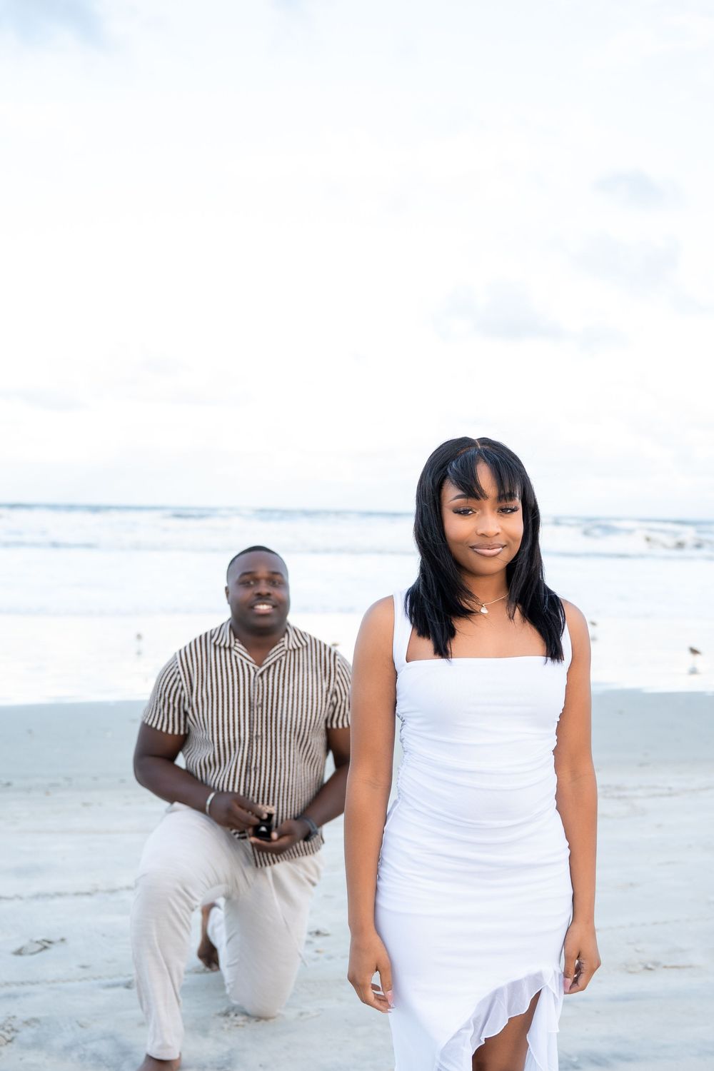 Man kneeling on beach with ring box proposing to woman in white dress by the ocean.
