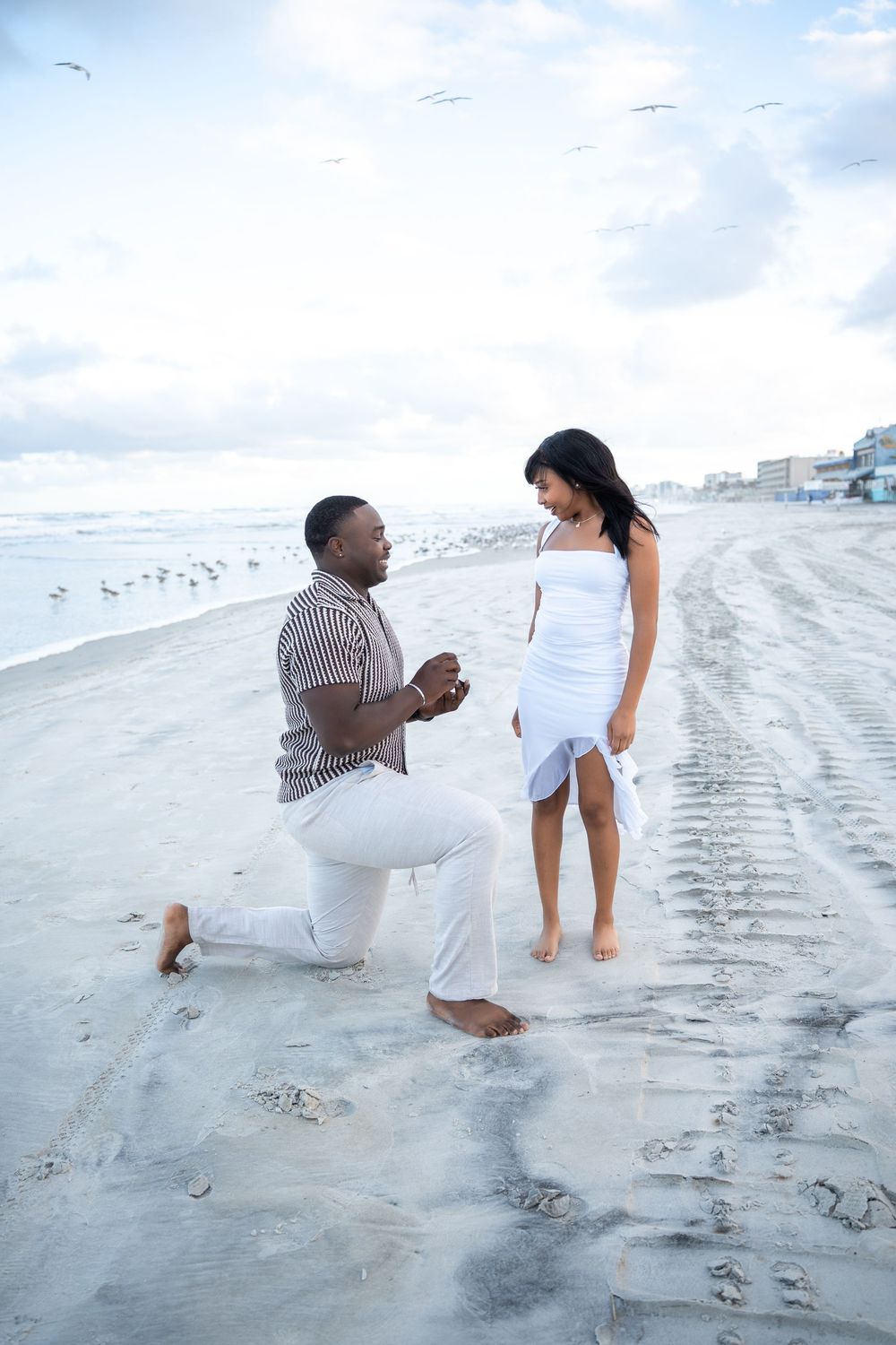 Man proposing on one knee to woman in white dress on sandy beach with ocean waves and seagulls in background.
