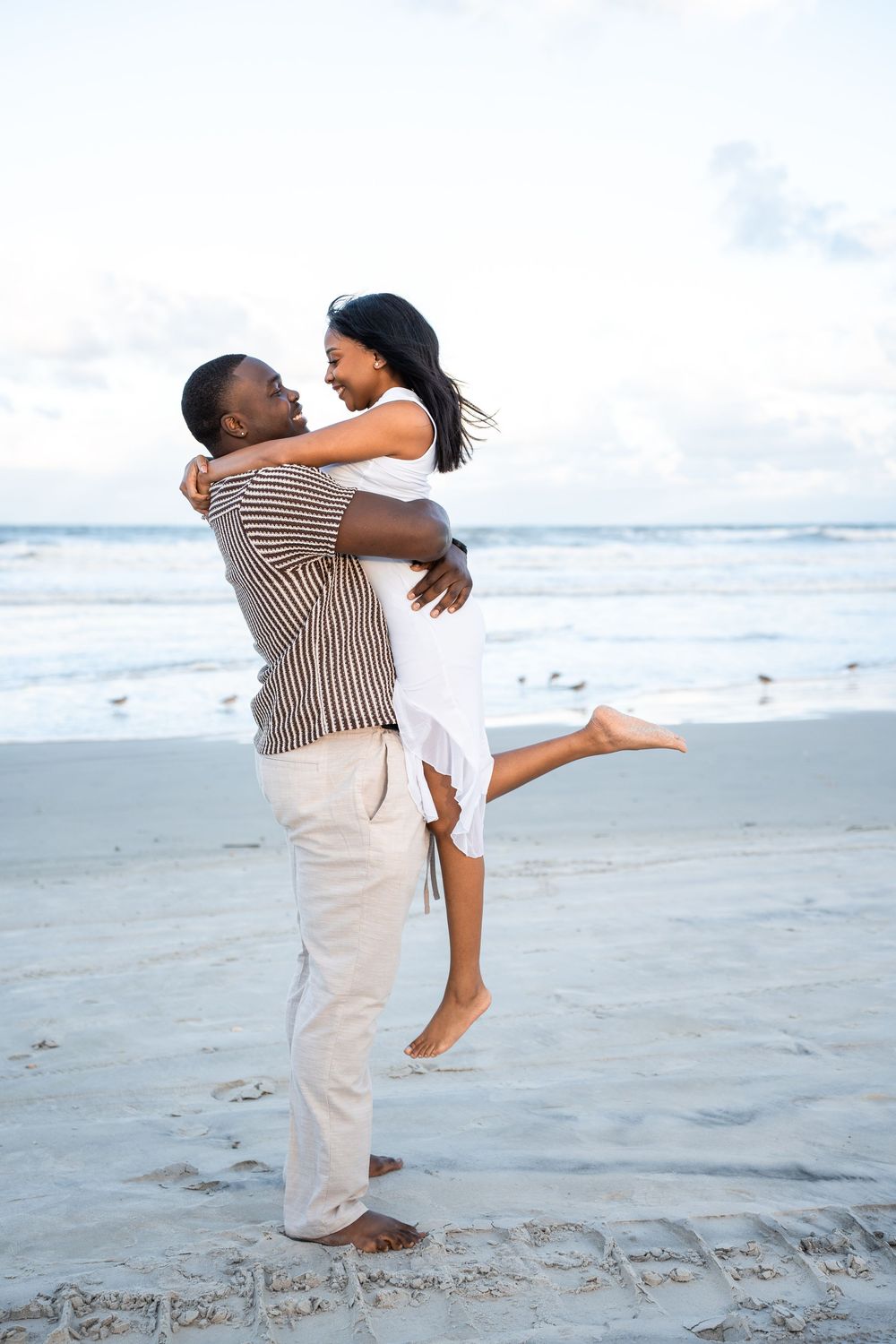 A joyful couple embraces on a sandy beach, man lifting woman in white dress, ocean waves in the background.