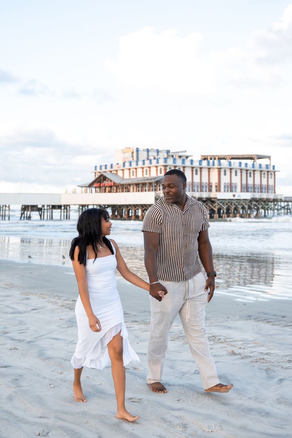 A couple holds hands walking barefoot on a sandy beach with a pier in the background at dusk.