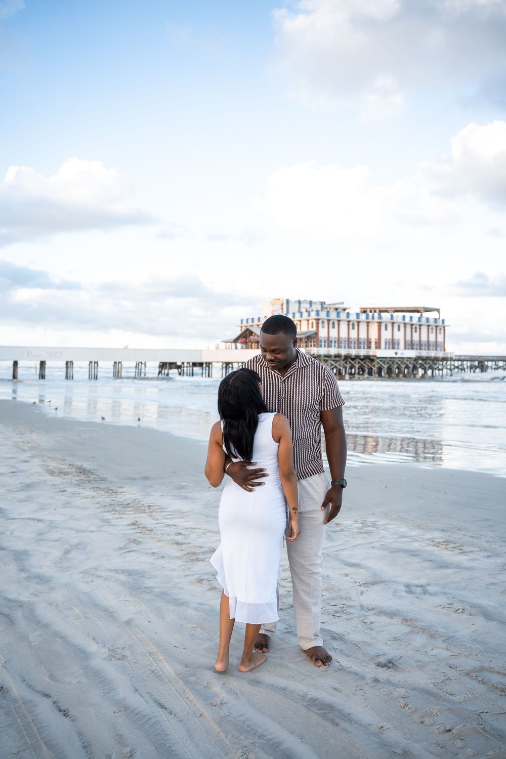 Couple walking barefoot on sandy beach near pier, man in striped shirt with woman in white dress.
