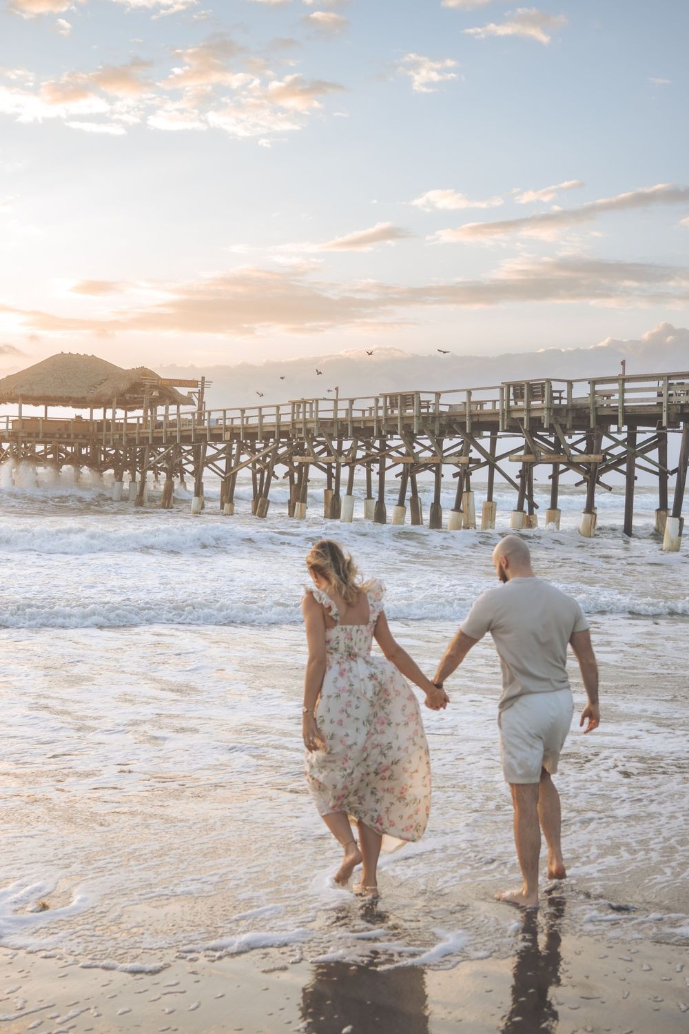 Couple holding hands walking barefoot on a sandy beach at sunset near a wooden pier with waves washing ashore.