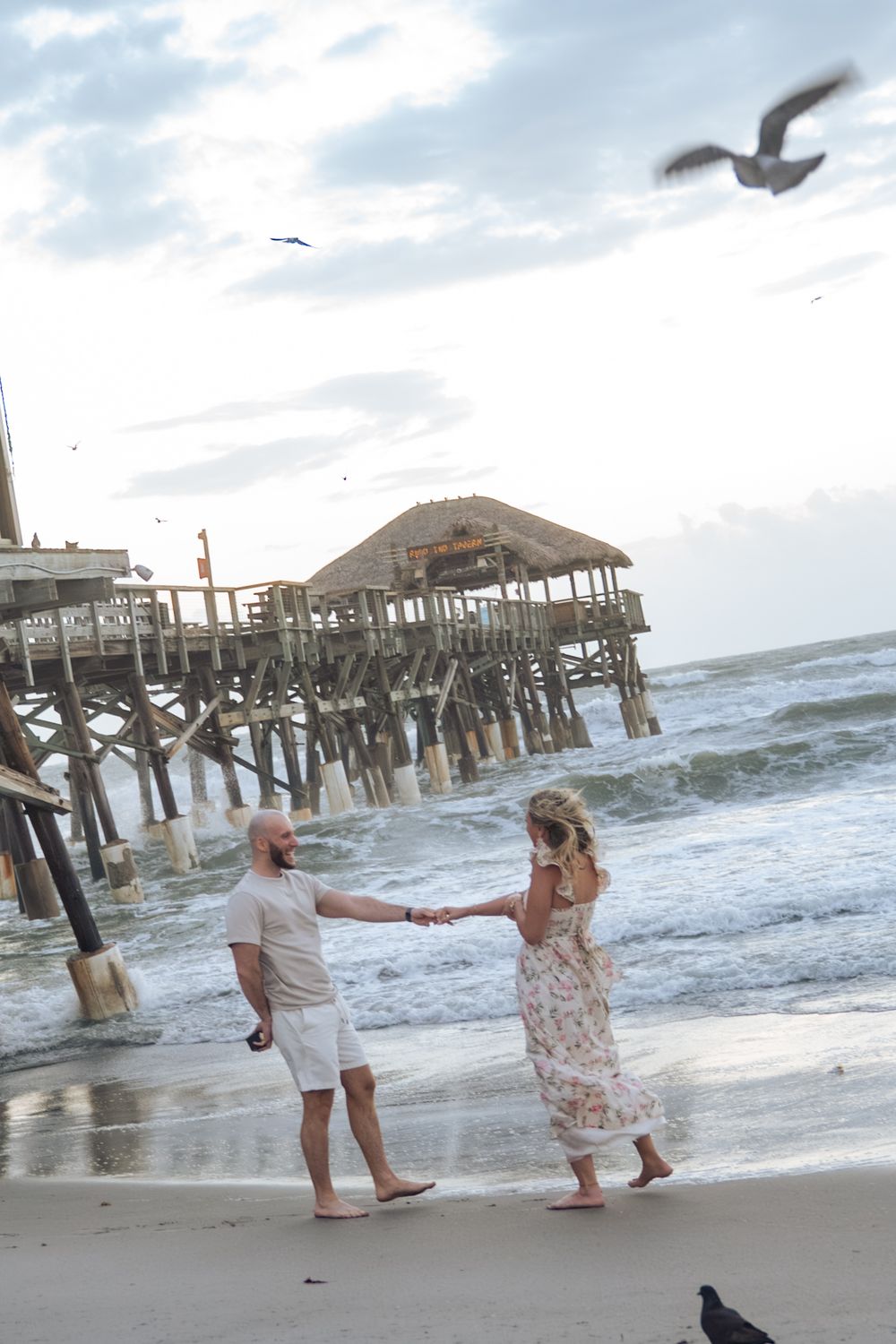 Couple holding hands on a sandy beach near a wooden pier with ocean waves and seagulls flying overhead.