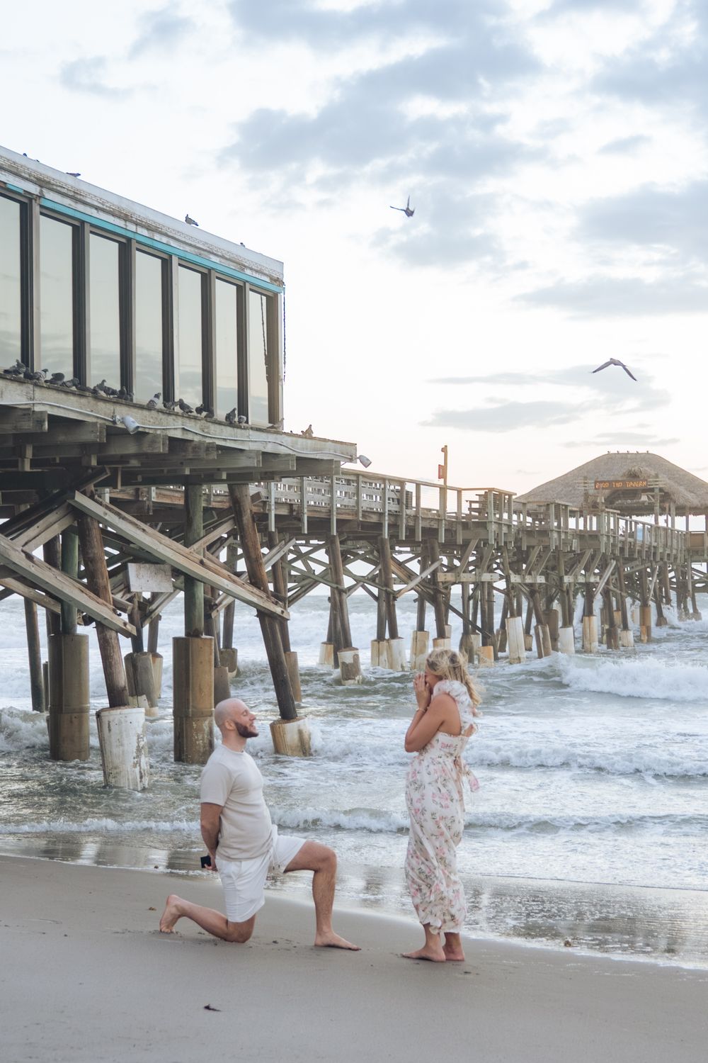 Man proposing on one knee to woman on beach near wooden pier with waves and seagulls in background.