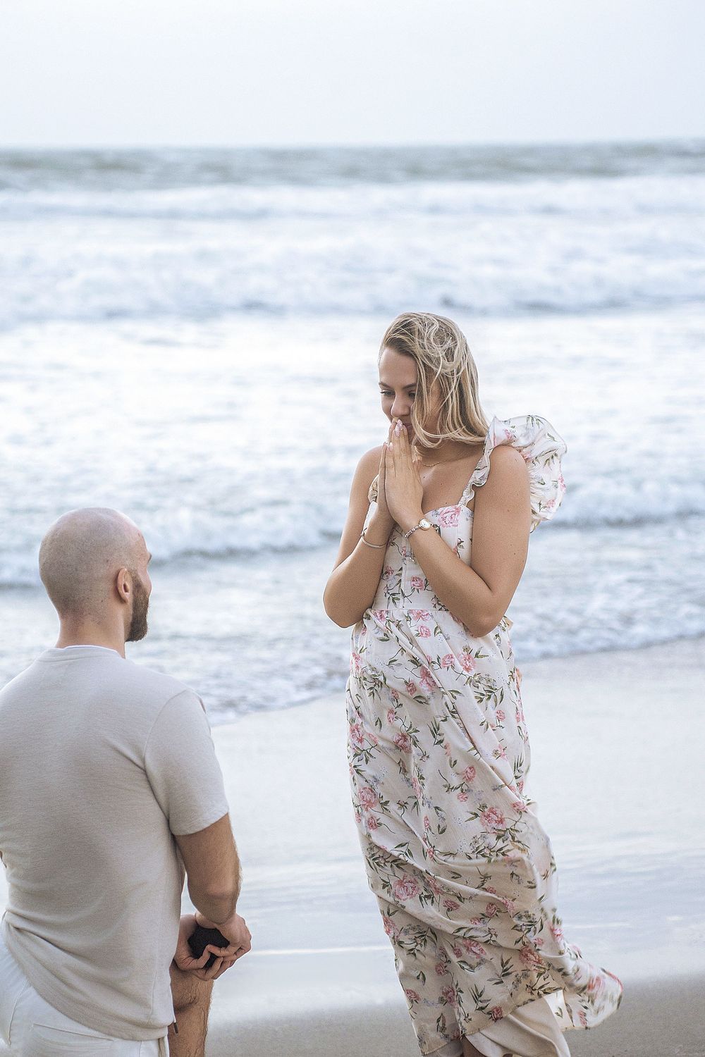 Man kneeling on beach proposing to surprised woman in floral dress with ocean waves in background.