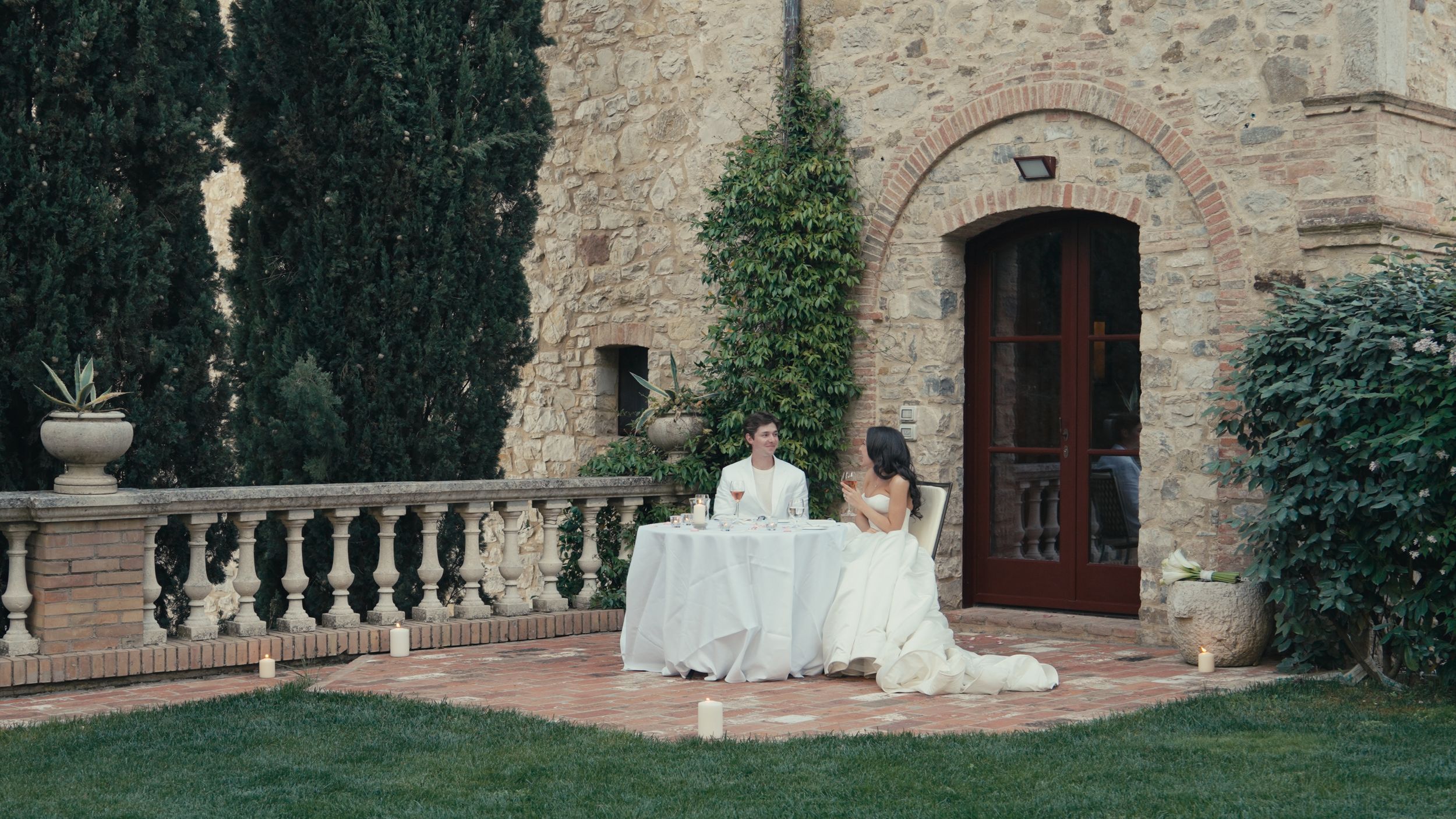 Couple shares an intimate outdoor dinner at a white-draped table beside a rustic stone villa surrounded by cypress trees.