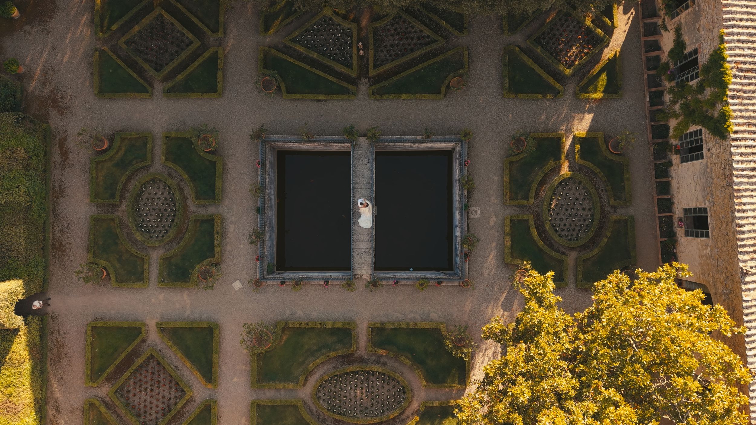 Aerial view of a formal garden with geometric patterns, twin rectangular pools, and golden autumn trees.