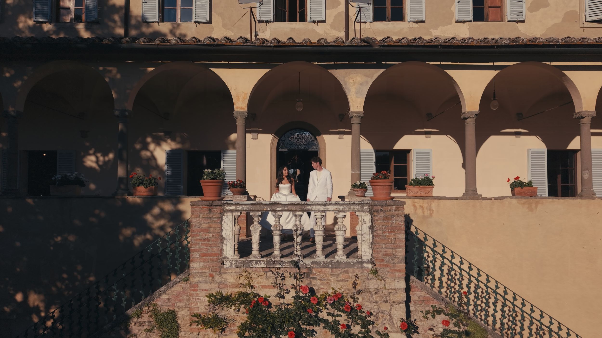 A couple stands on a sunlit terrace of a Tuscan villa with arched colonnades and terracotta brick details.