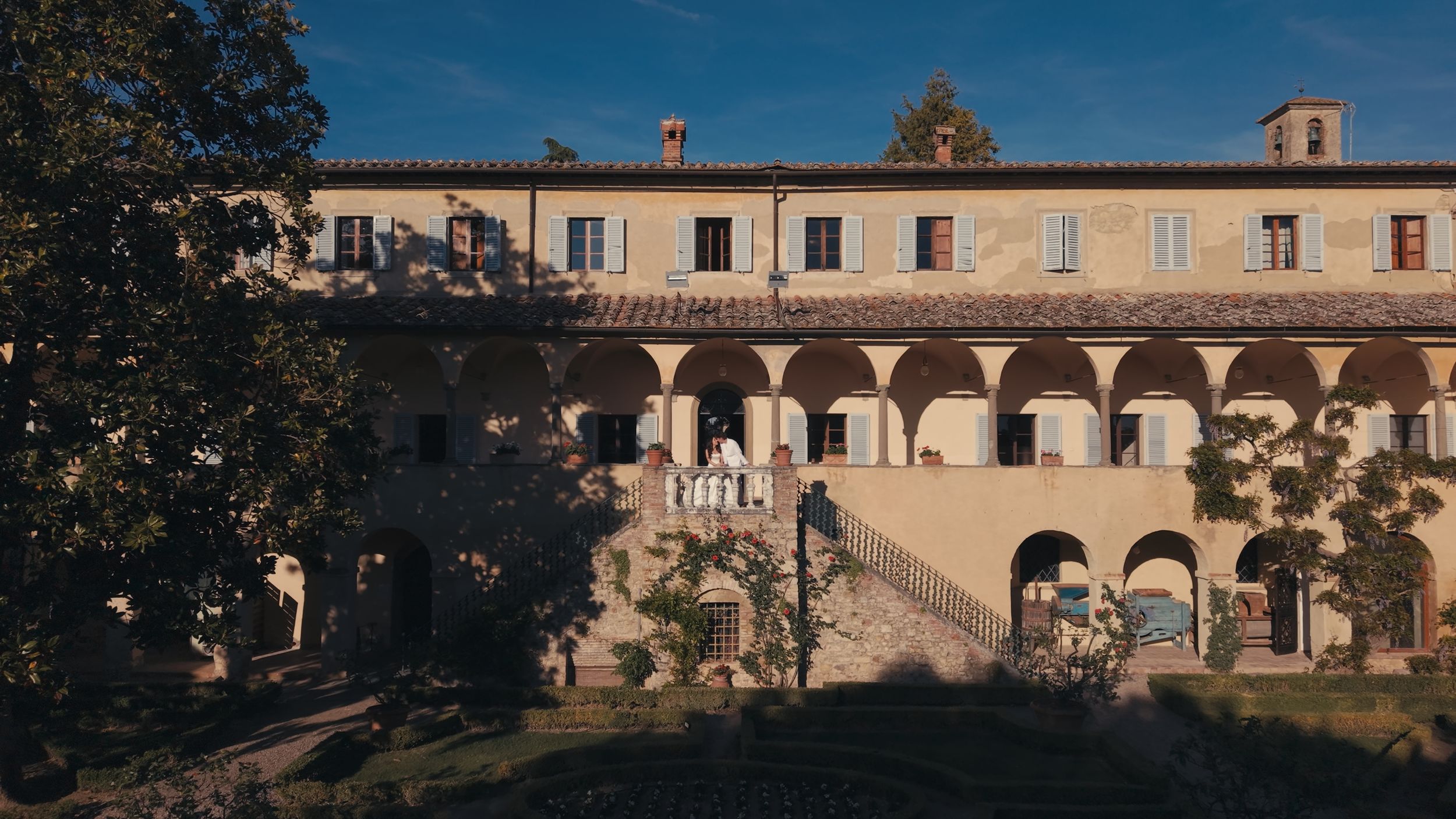 Historic Italian villa with arched colonnade facade, lush trees, and warm sunlight in a serene countryside setting.
