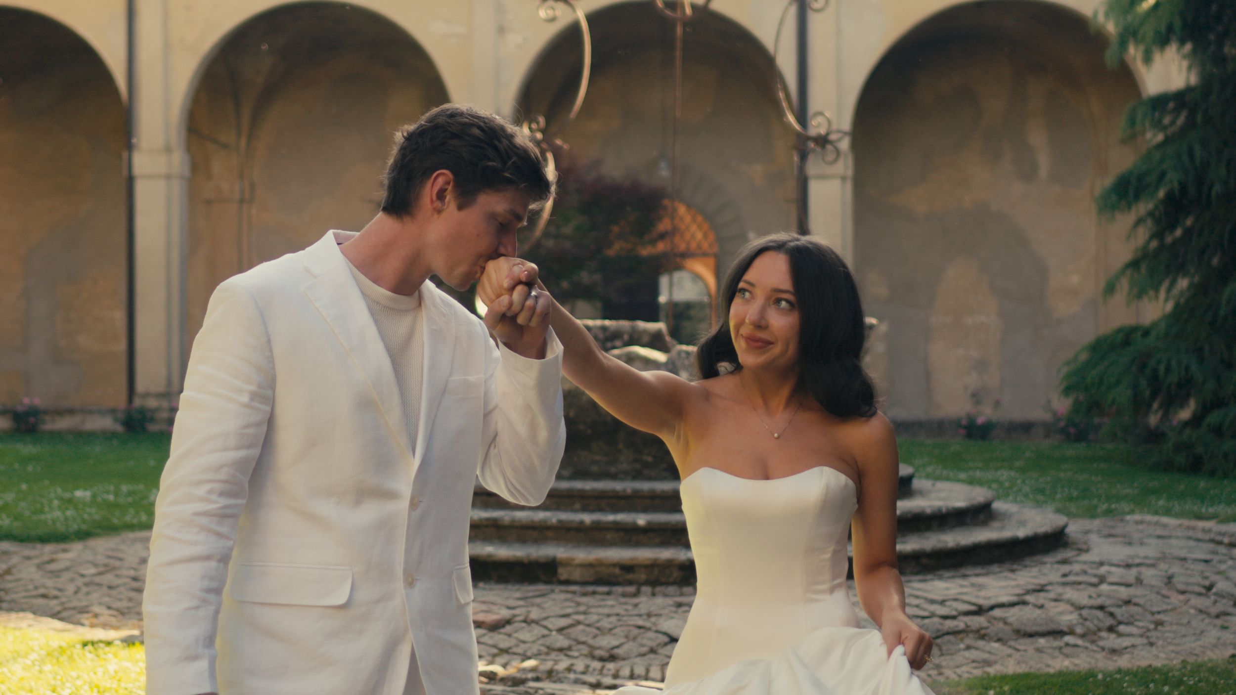 Elegant couple in wedding attire sharing a romantic moment in a sunlit courtyard, groom in white suit kissing bride's hand.
