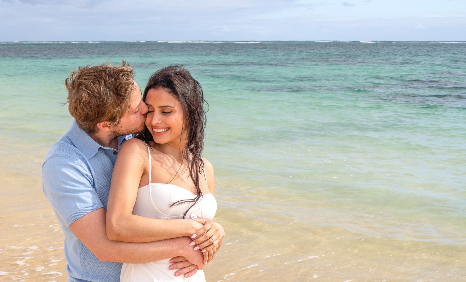 A romantic couple embraces on a tropical beach, man kissing woman's cheek with turquoise ocean waves behind them.