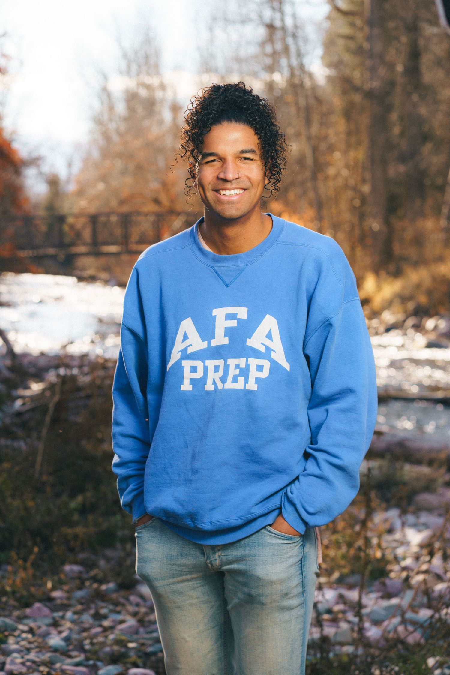 Smiling young man wearing a blue AFA Prep crewneck sweatshirt and jeans, standing outdoors near a creek.