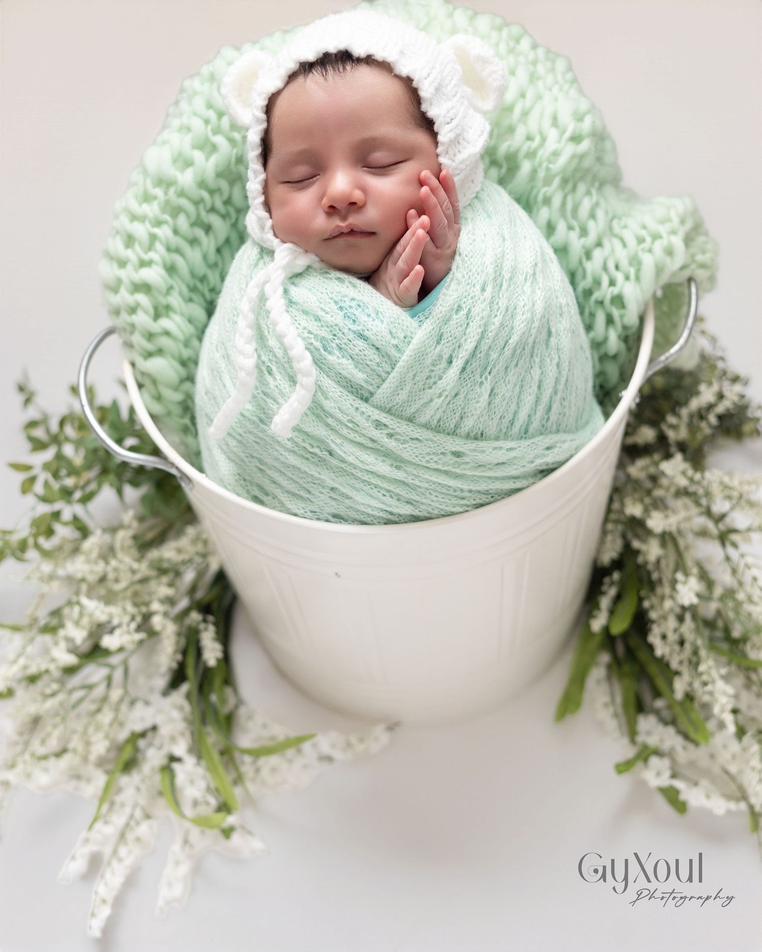 Newborn baby wrapped in mint green knit swaddle, wearing white bear bonnet, nestled in white bucket with flowers.