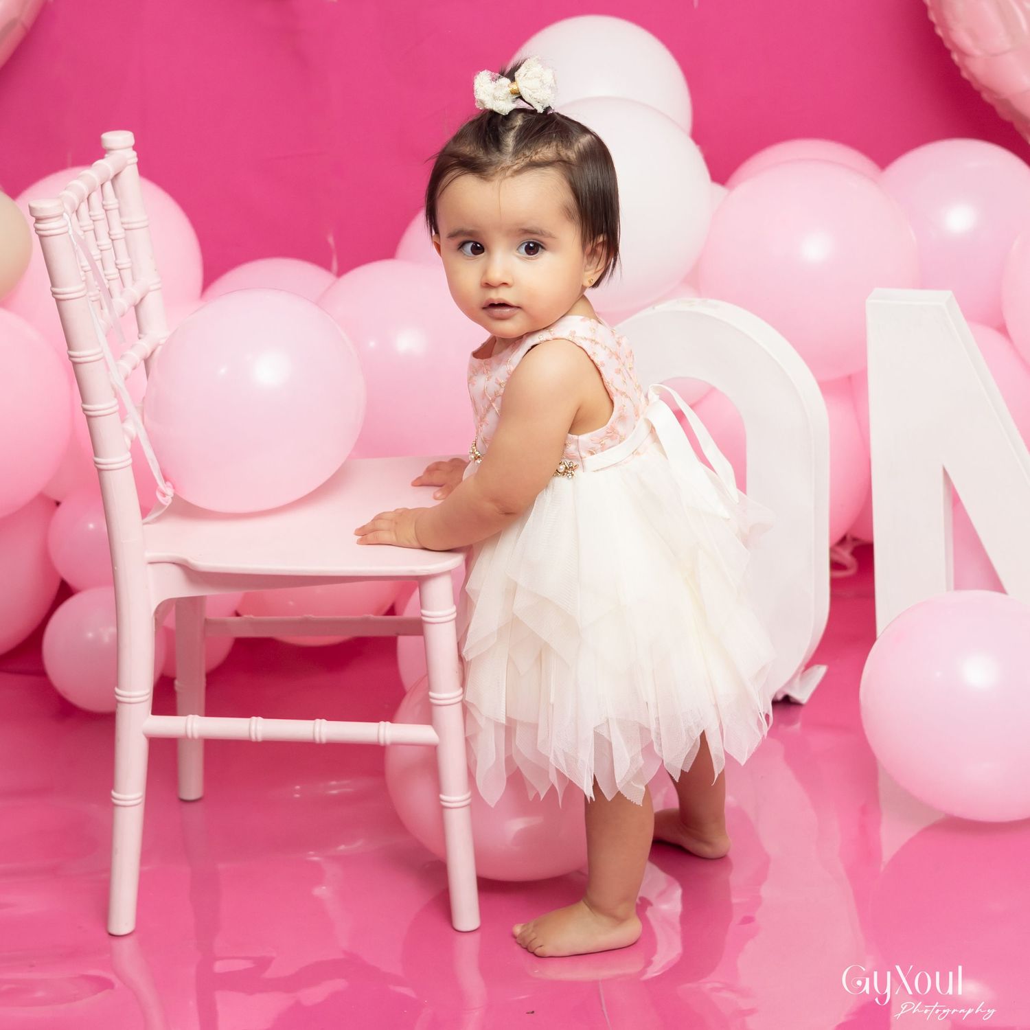 Baby girl in white tutu dress standing by small white chair with pink balloons for first birthday photo.