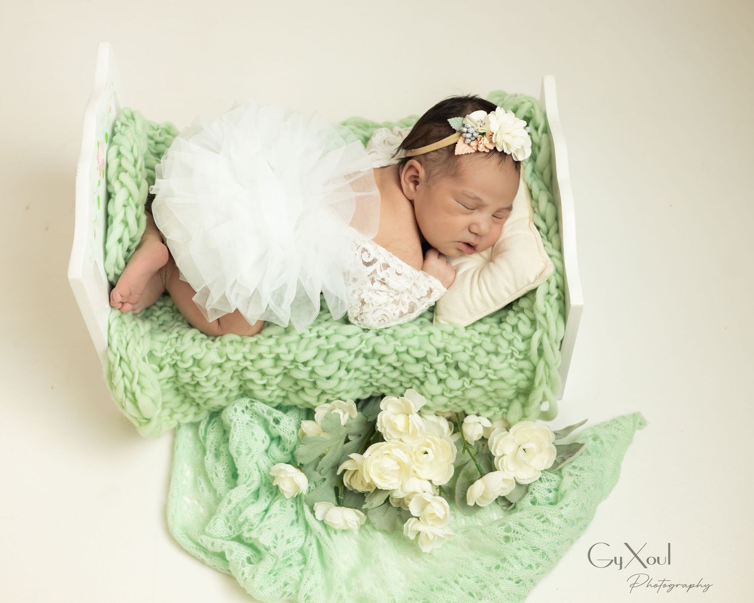 Newborn baby sleeping in a mint green knit basket wearing a white tutu and floral headband with white roses.