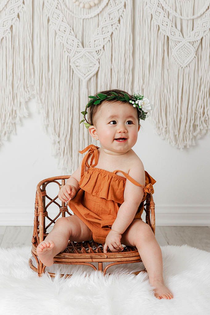 Happy baby in rust romper with green floral crown sits on rattan chair with macrame backdrop.