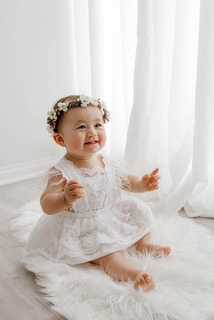 Smiling baby girl in white lace dress and floral crown sits on fluffy rug near sheer curtains.