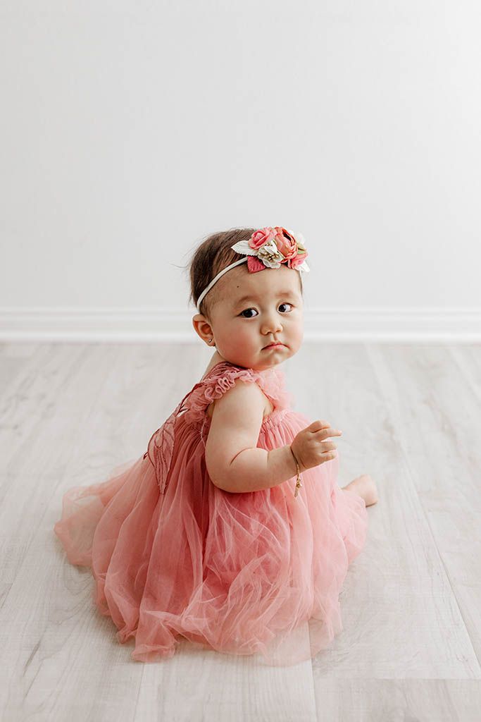 Baby girl in pink tulle dress and floral headband sitting on light wood floor.