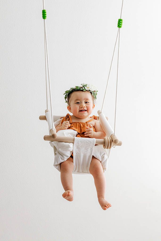Smiling baby in orange romper and flower crown sitting in a hanging wooden swing.