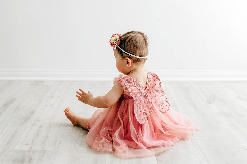 Baby girl in pink tutu dress with butterfly wings and floral headband sitting on hardwood floor.