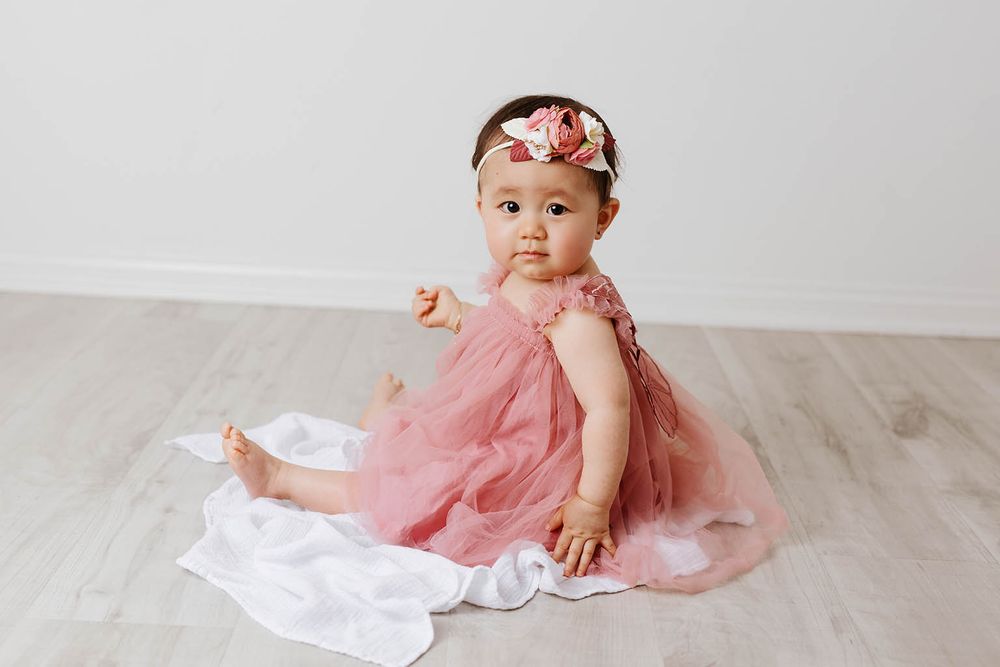 Baby girl in pink tulle dress sitting on white blanket on hardwood floor.