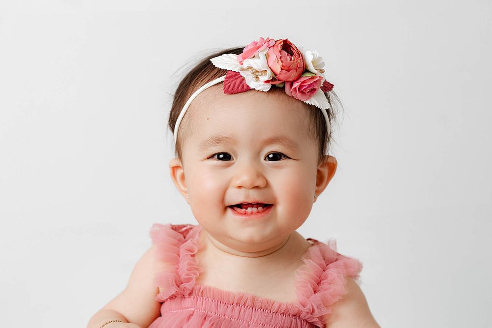 Smiling baby girl wearing a pink ruffle outfit and floral headband poses for a portrait photo.
