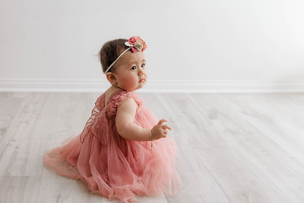 Baby girl in pink tulle dress with floral headband sitting on hardwood floor.