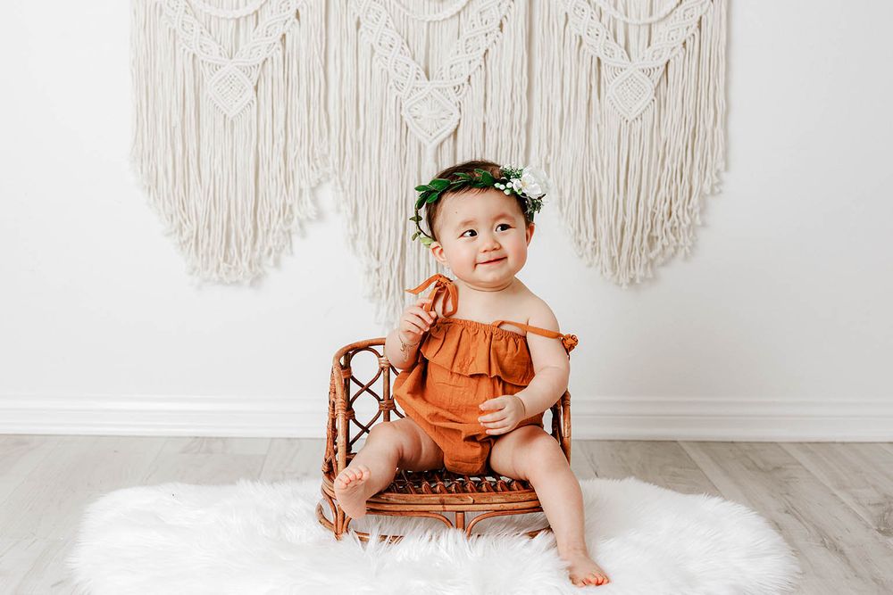 Baby girl in rust romper with green floral crown sitting in wicker chair on white fur rug.