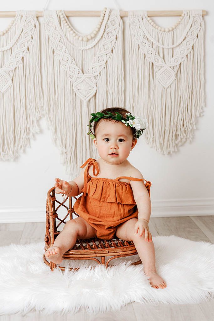 Cute baby in rust romper with floral crown sitting on wicker bench with macrame backdrop.