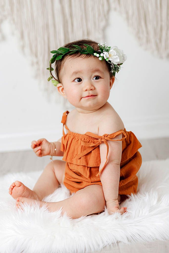 Baby girl in rust orange romper with green and white floral crown sitting on fur rug.