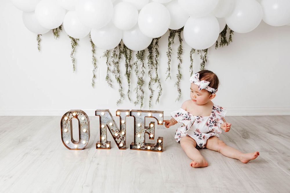 Baby girl in floral romper sitting next to illuminated ONE marquee letters with balloons.