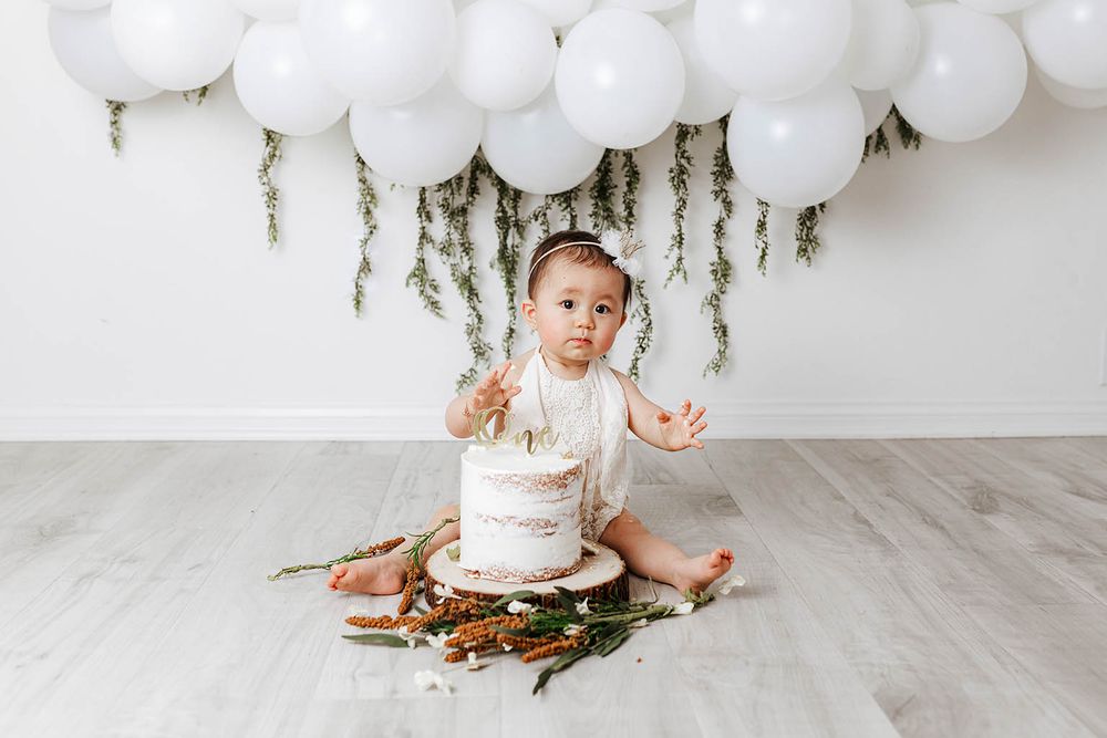 Baby smashing white first birthday cake surrounded by balloons and greenery.