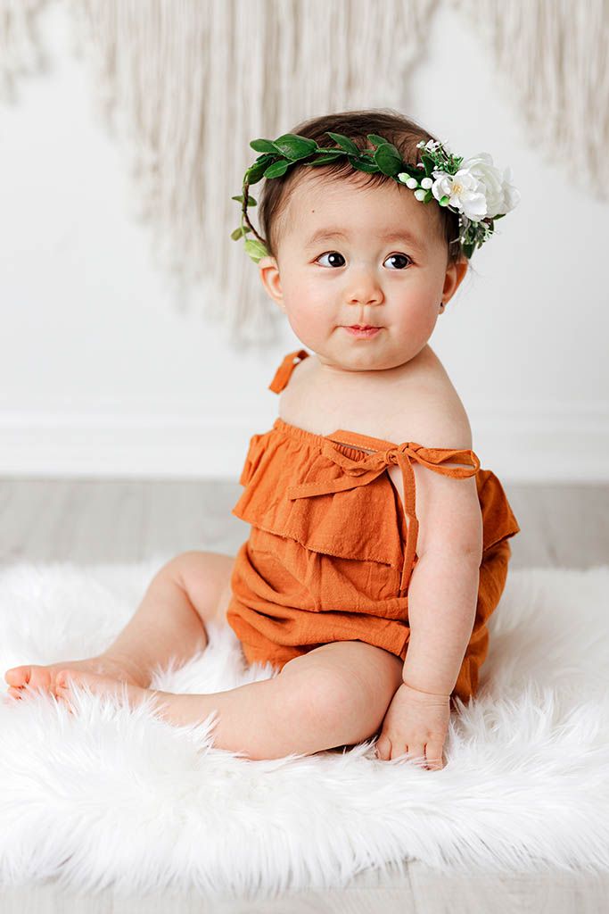 Adorable baby in rust orange romper with white floral green leaf crown on fur rug.