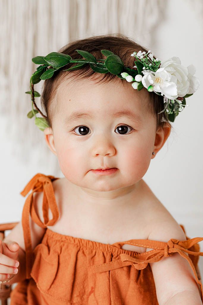 Close-up of baby in orange romper wearing white flower and greenery crown headband.