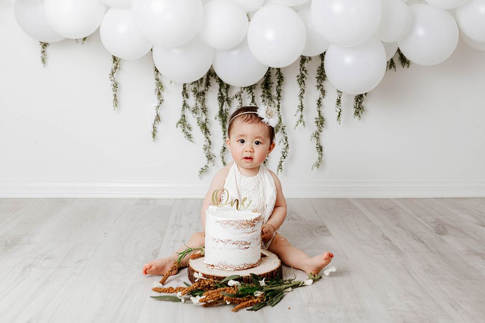 Baby girl sitting with white first birthday cake, white balloons and greenery backdrop.