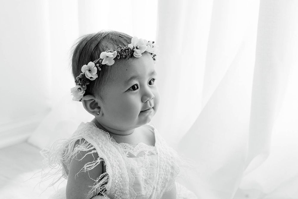 Black and white portrait of baby in lace dress with floral headband by sheer curtains.