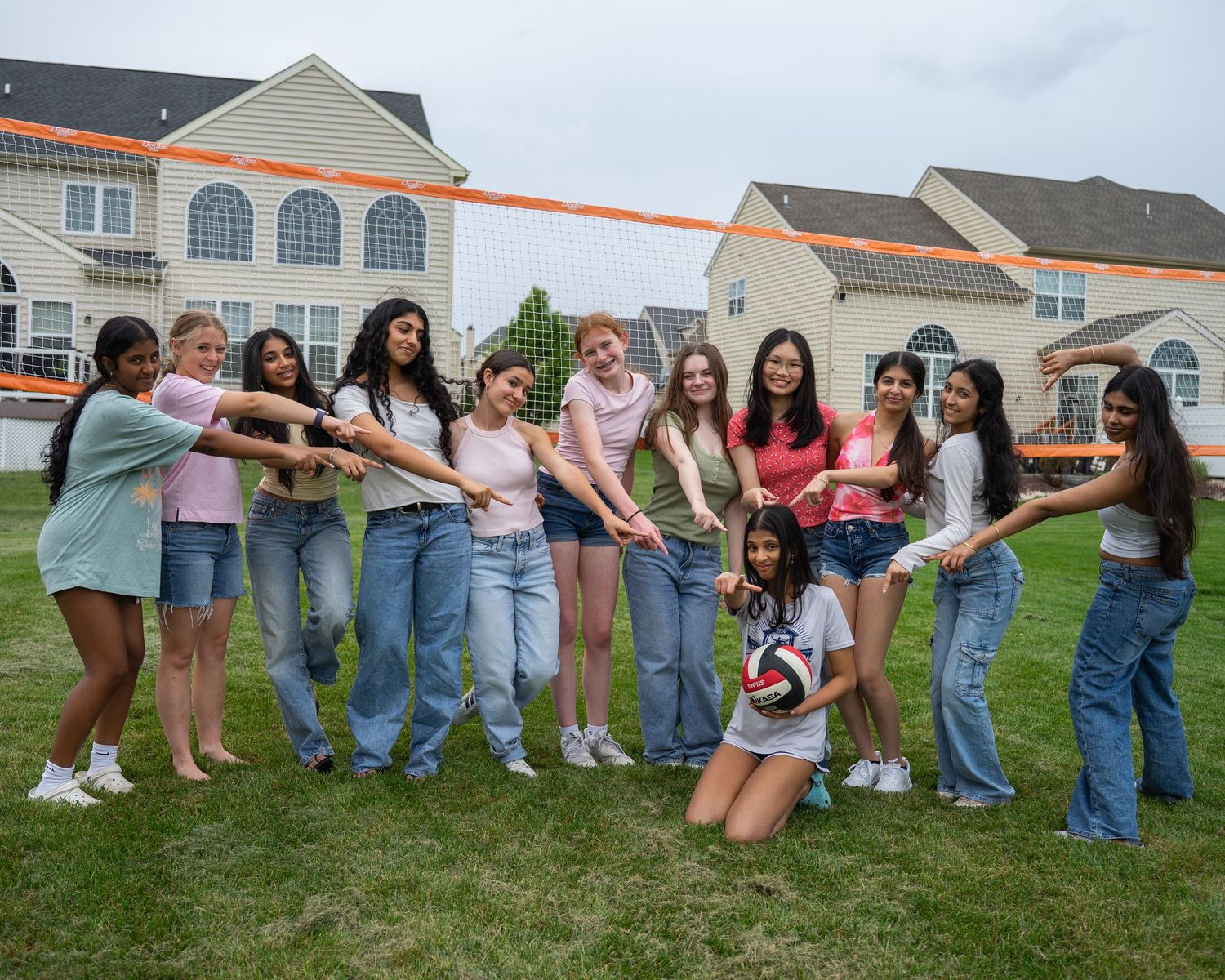 Group of teenage girls posing together outdoors near a volleyball net on a green lawn with houses in the background.
