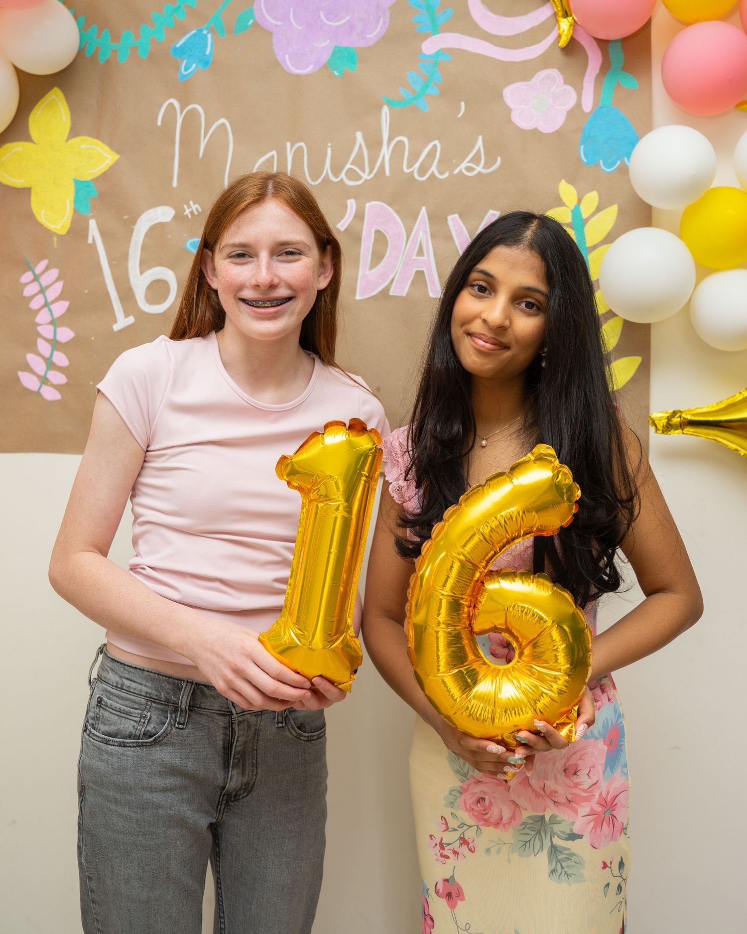 Two girls holding gold number 16 balloons at Manisha's 16th birthday party with floral balloon backdrop.