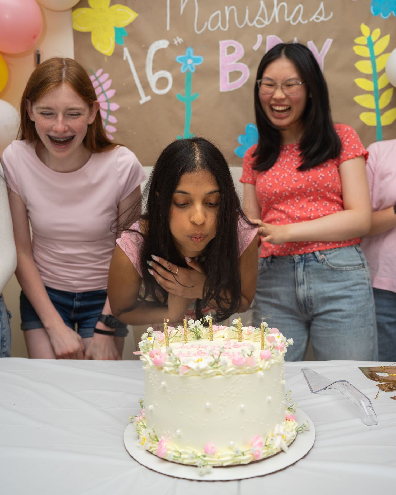 Teen girl blows out candles on floral birthday cake at Manisha's 16th birthday party with friends laughing behind her.