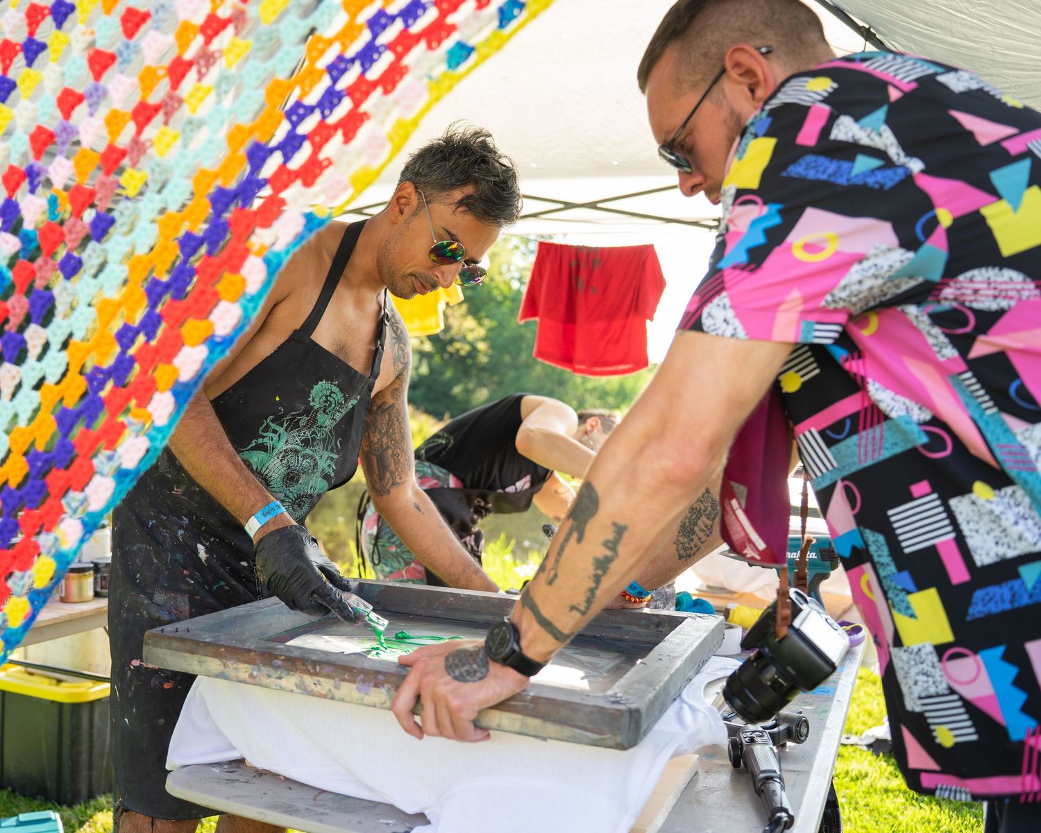 Two artists screen printing on fabric under a colorful decorative canopy at an outdoor festival event.