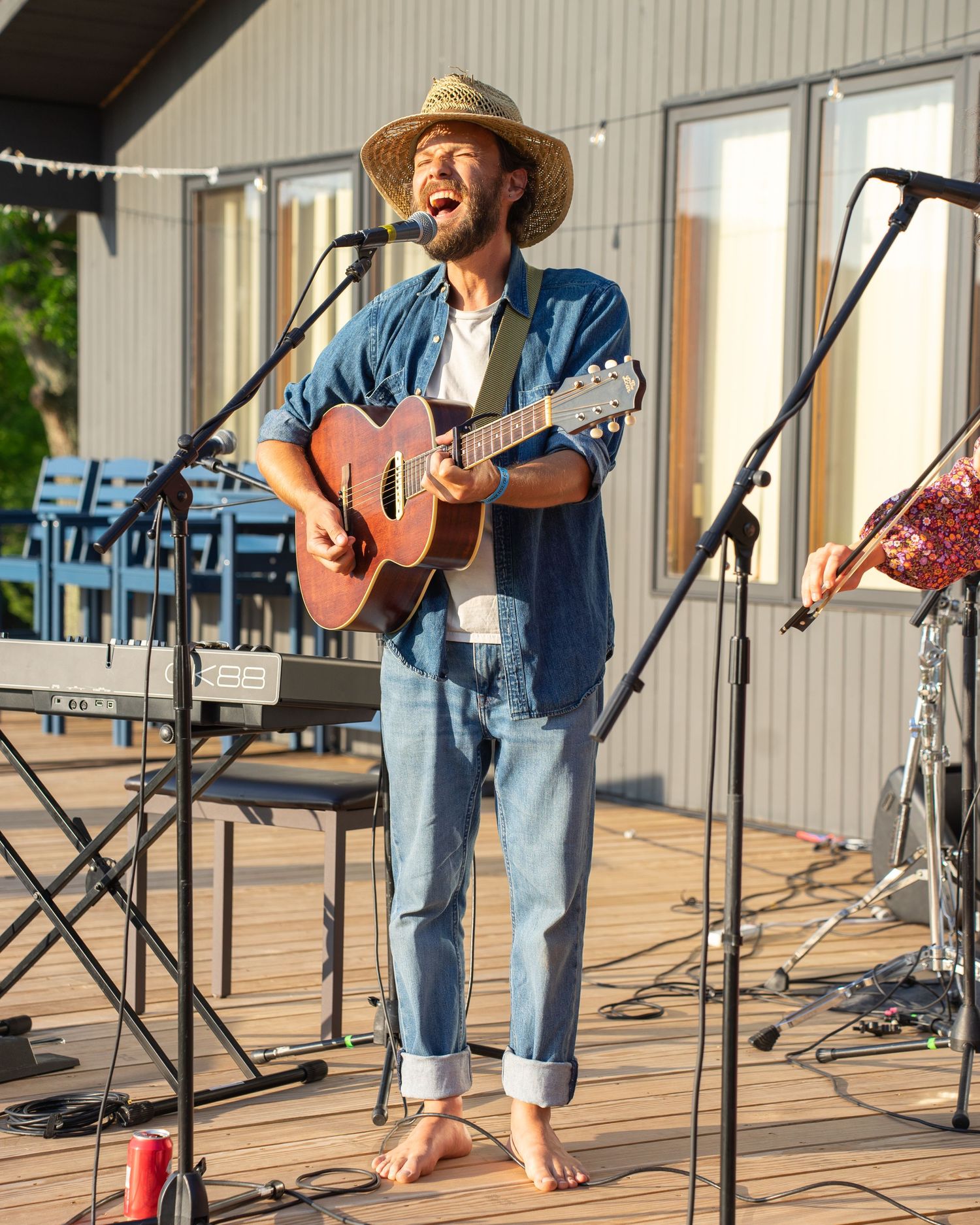 Barefoot male musician playing acoustic guitar and singing outdoors on a wooden deck stage wearing a straw hat.