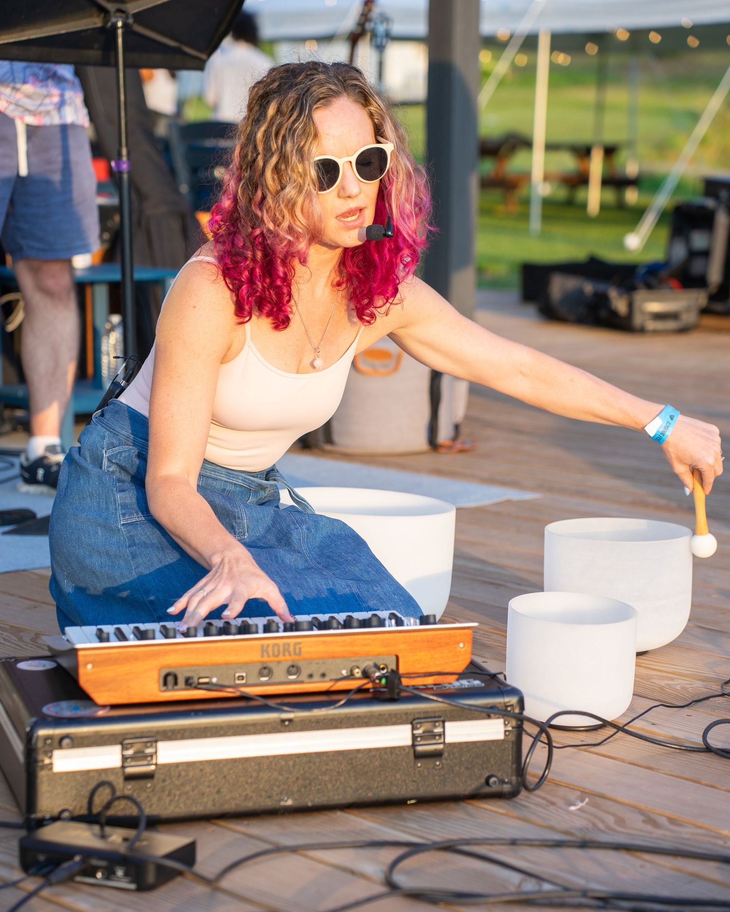 Woman with red curly hair plays crystal singing bowls and keyboard outdoors at a music event.