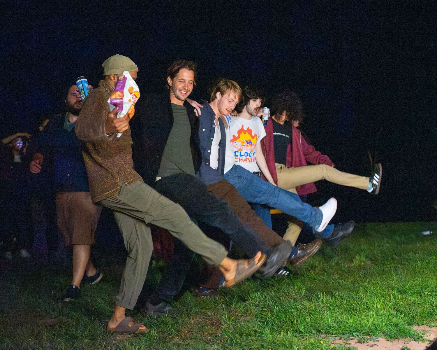 Group of young friends posing playfully outdoors at night, kicking legs up together in a fun candid photo.