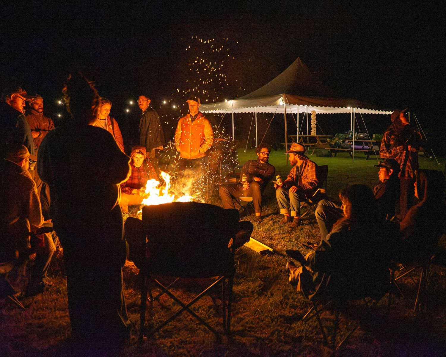 Group of people gathered around a glowing campfire at night with a lit tent canopy in the background.