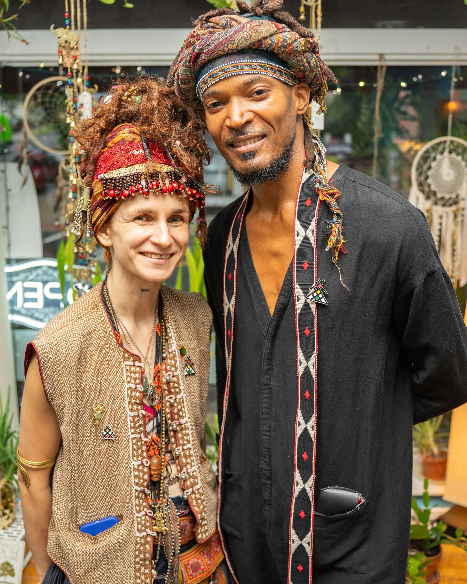 Two people in colorful bohemian headwear and ethnic clothing pose together at an outdoor market.