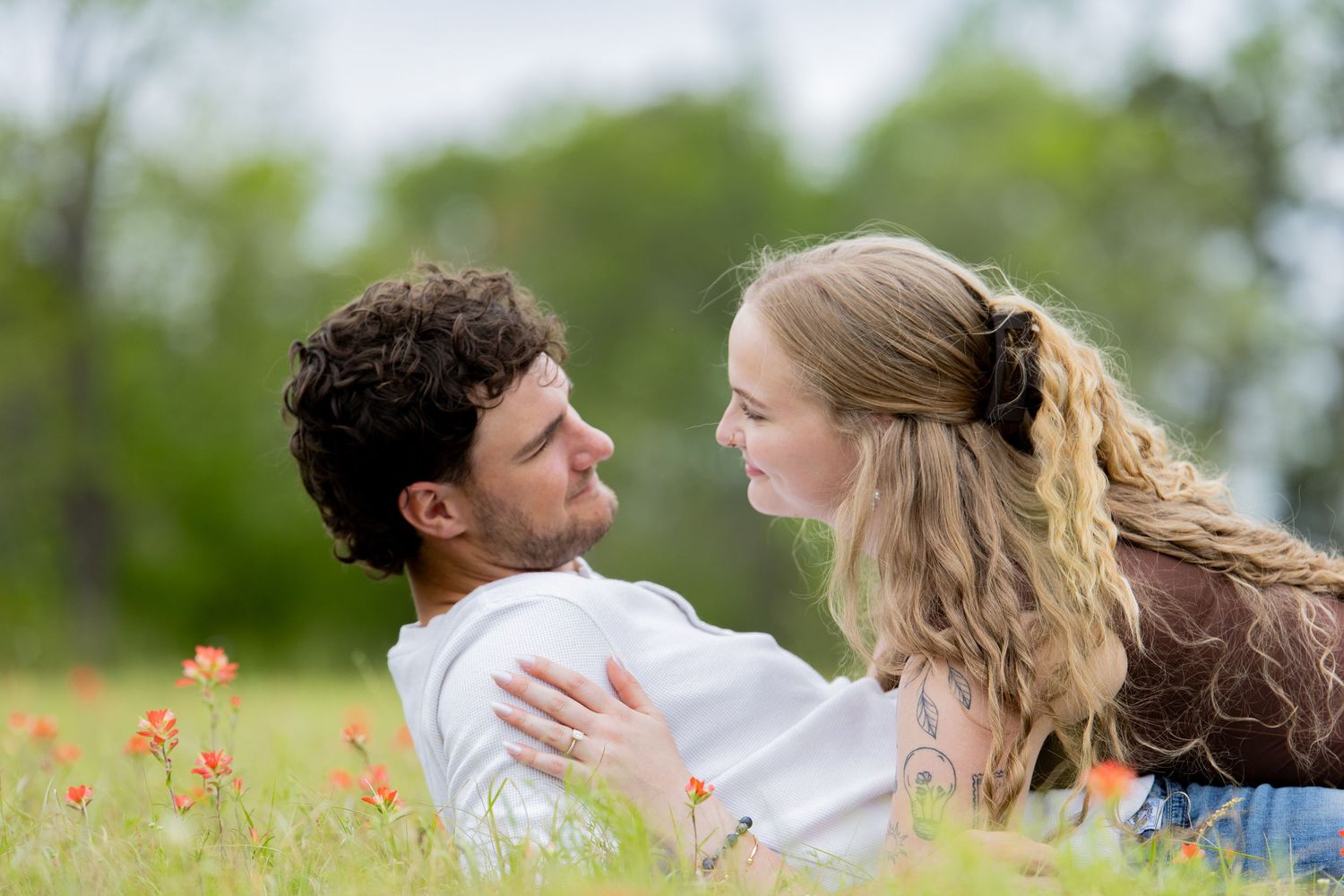Couple poses back-to-back outdoors, woman in white dress with tattoos smiles, man in striped shirt and jeans looks away.