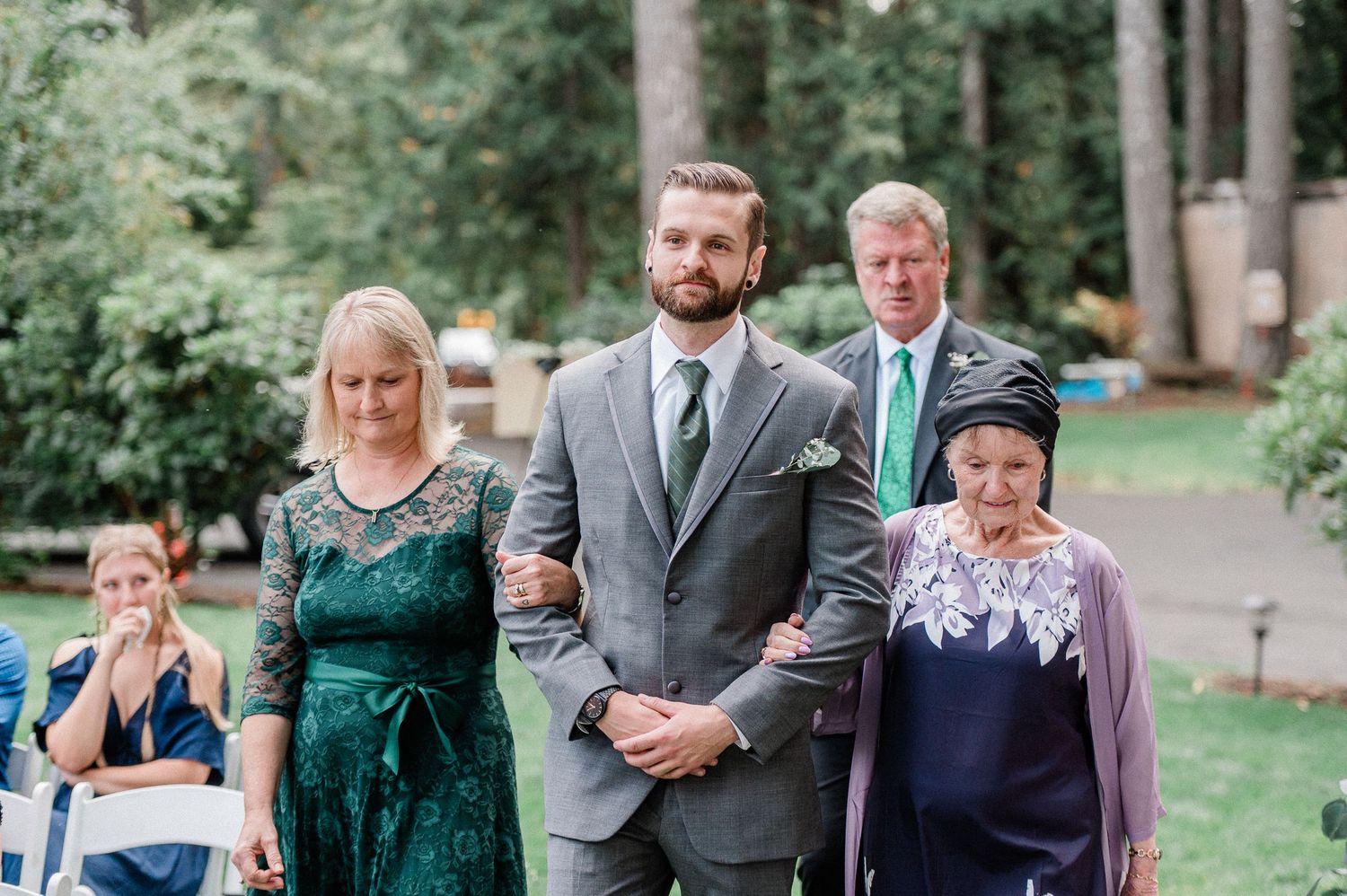 Groom walks down outdoor wedding aisle escorted by two women in elegant dresses, with a man in a suit behind them.