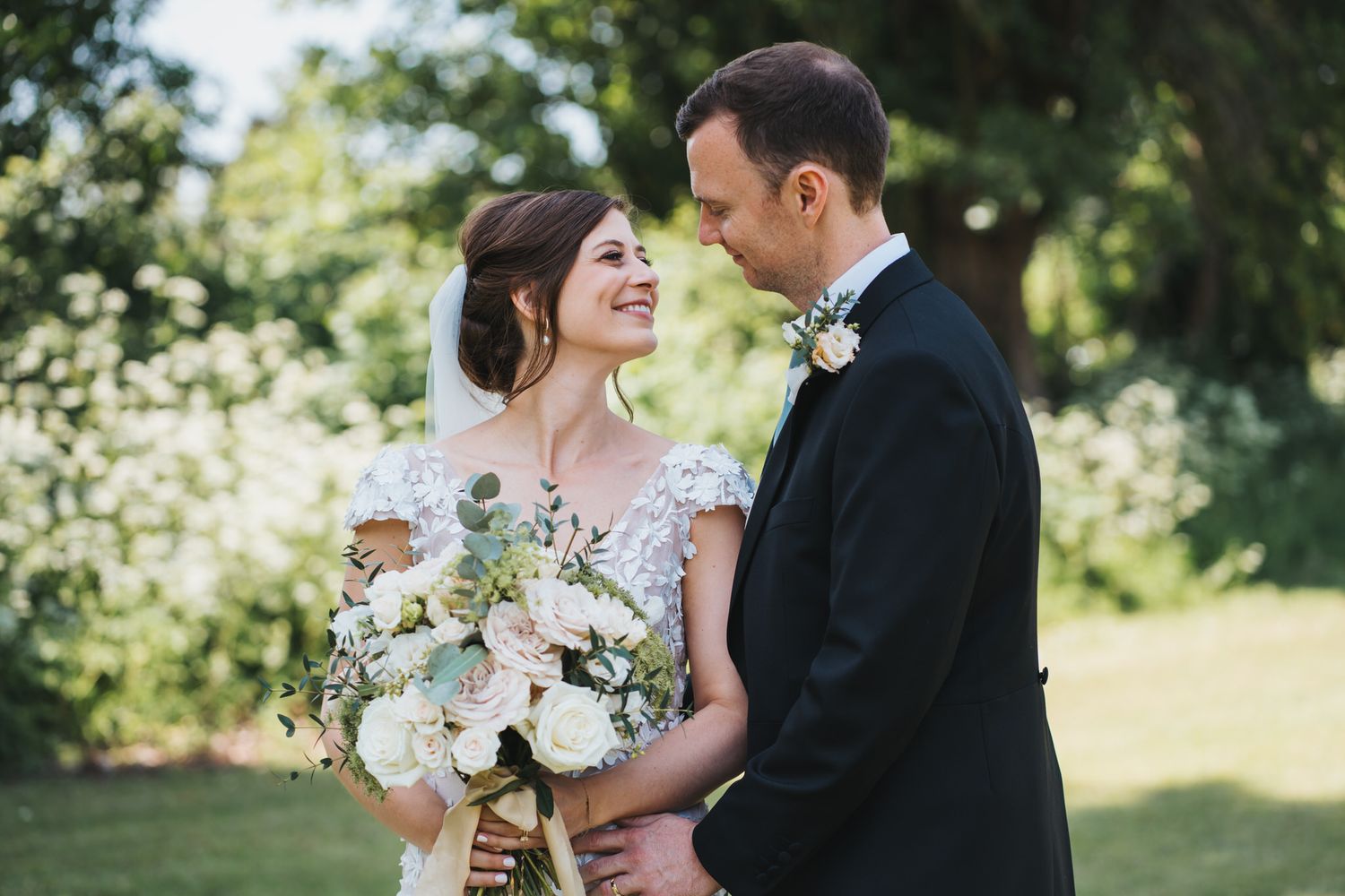 Bride and groom smiling at each other outdoors, bride holding white and blush floral bouquet, groom in black tuxedo.