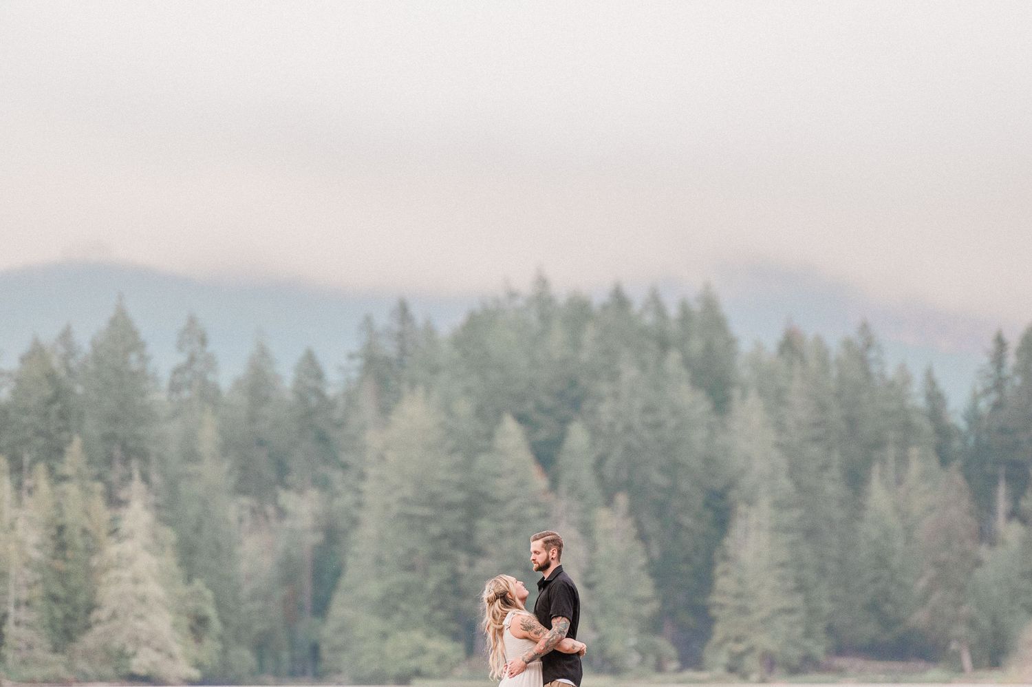 Couple embracing in a misty forest landscape with tall evergreen trees and mountain backdrop.